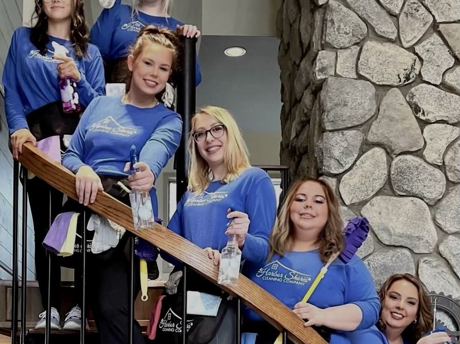 Team of Harbor Shores Cleaning Company members in blue shirts, holding cleaning supplies on staircase, showcasing professional cleaning services for homes and businesses in Northern Michigan.